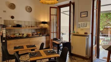 A cozy dining area with wooden tables and chairs, a kitchen with shelves and jars, and a view of greenery outside.