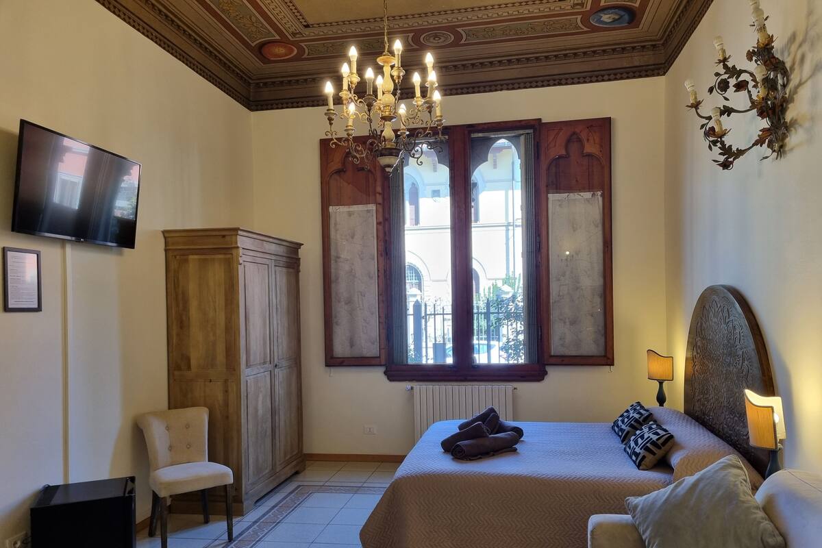 A bedroom with a large bed, a wooden wardrobe, a chair, a TV, and a chandelier.