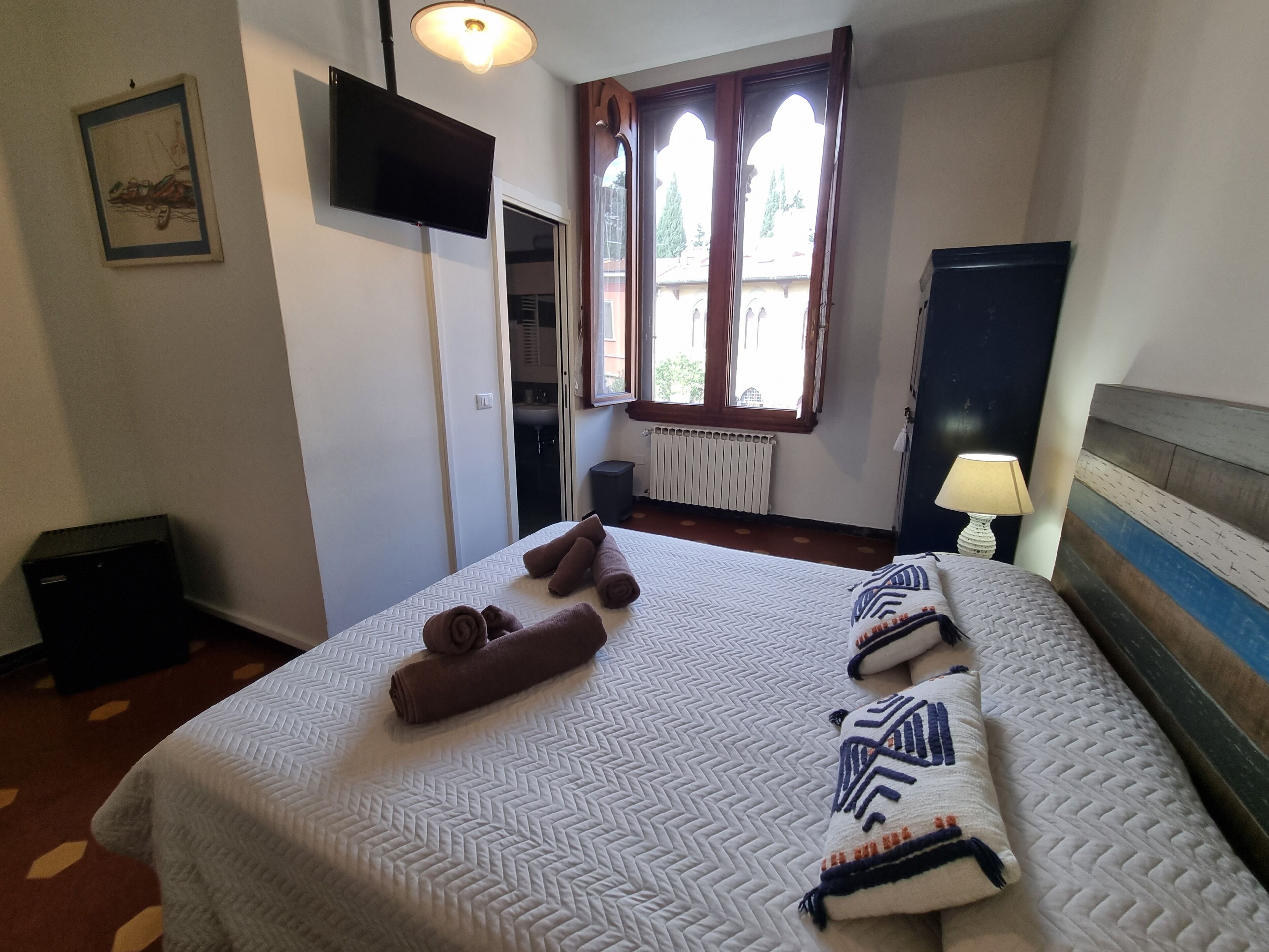 A hotel room with a bed, a TV mounted on the wall, a window with curtains, a radiator, and a bedside lamp.