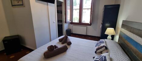 Et hotelværelse med seng, fjernsyn ophængt på væggen, et vindue med gardiner, en radiator og en sengetårnlampe.