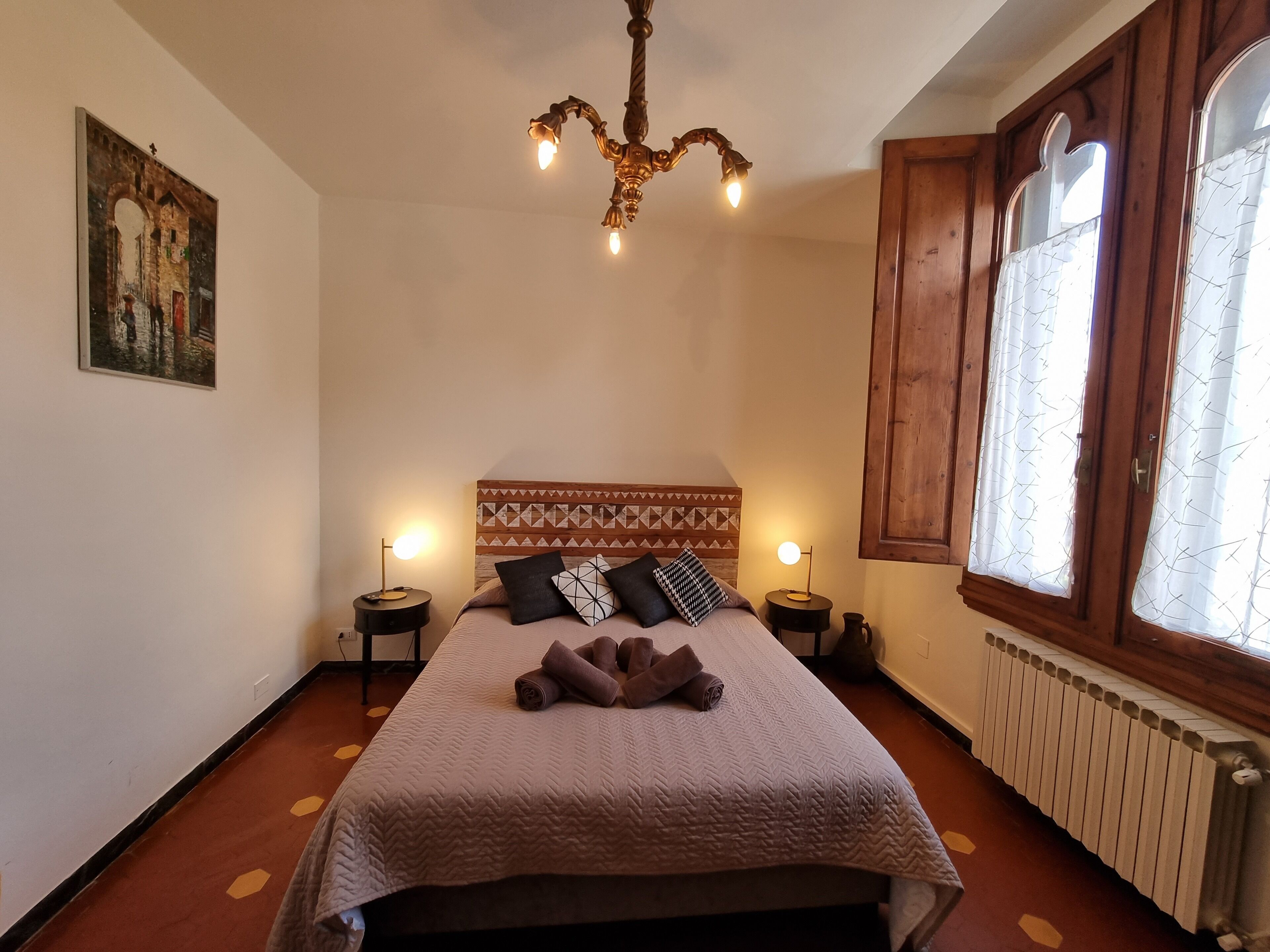 Ein Schlafzimmer mit einem Bett, zwei Nachttischen, einer Chandelierleuchtre, einem Fenster mit Vorhängen und einem Heizkörper.