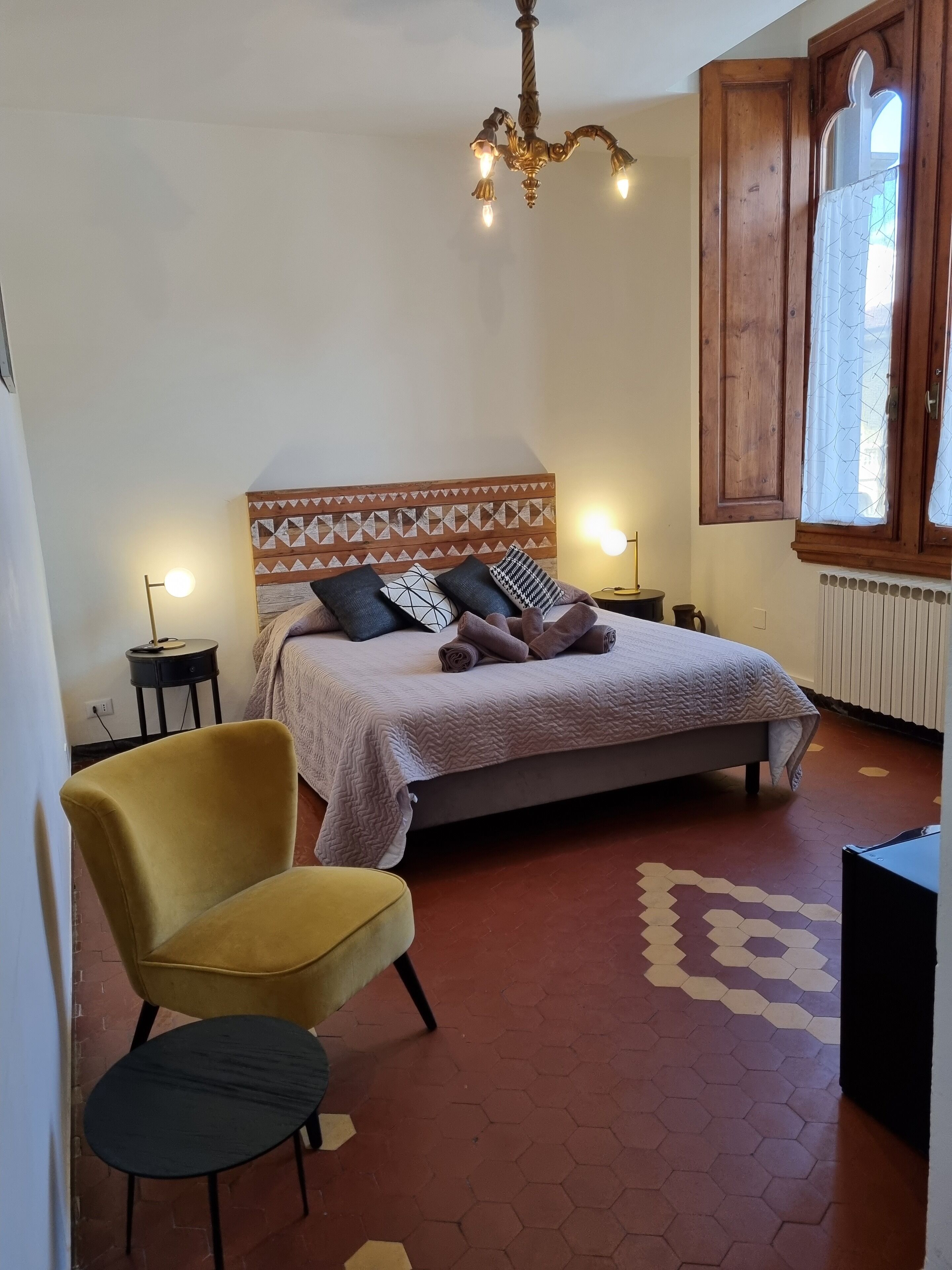 Un dormitorio con una cama, un sillón amarillo, una mesita, una araña y una ventana con cortinas.