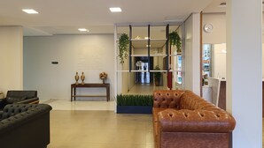 Um lobby moderno com um sofá de couro preto, um sofá de couro marrom, uma mesa consola com objetos decorativos e um separador de vidro com vista para outra área.