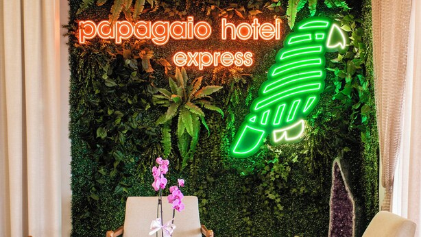 Um saguão de hotel com um letreiro de neon verde que diz "papagayo hotel express", uma parede coberta por plantas e duas poltronas com uma pequena mesa no centro.