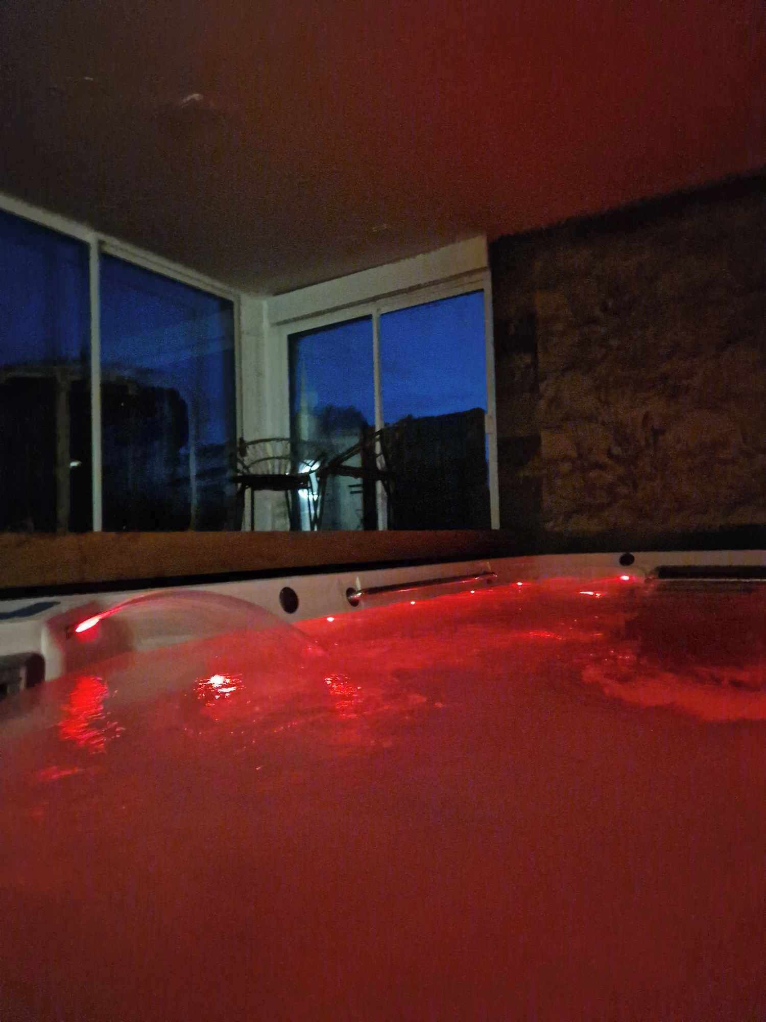 A hot tub with red lighting, a view of the outside through large windows, and a stone wall.