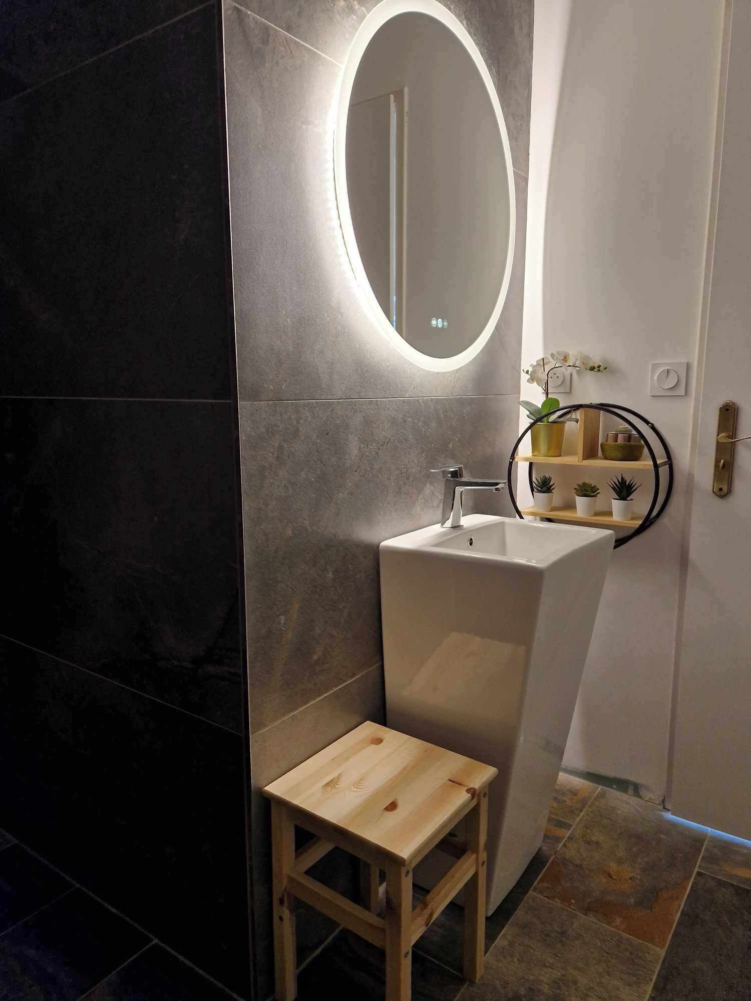 A modern bathroom with a white pedestal sink, a wooden stool, a round mirror, and a small shelf with plants.