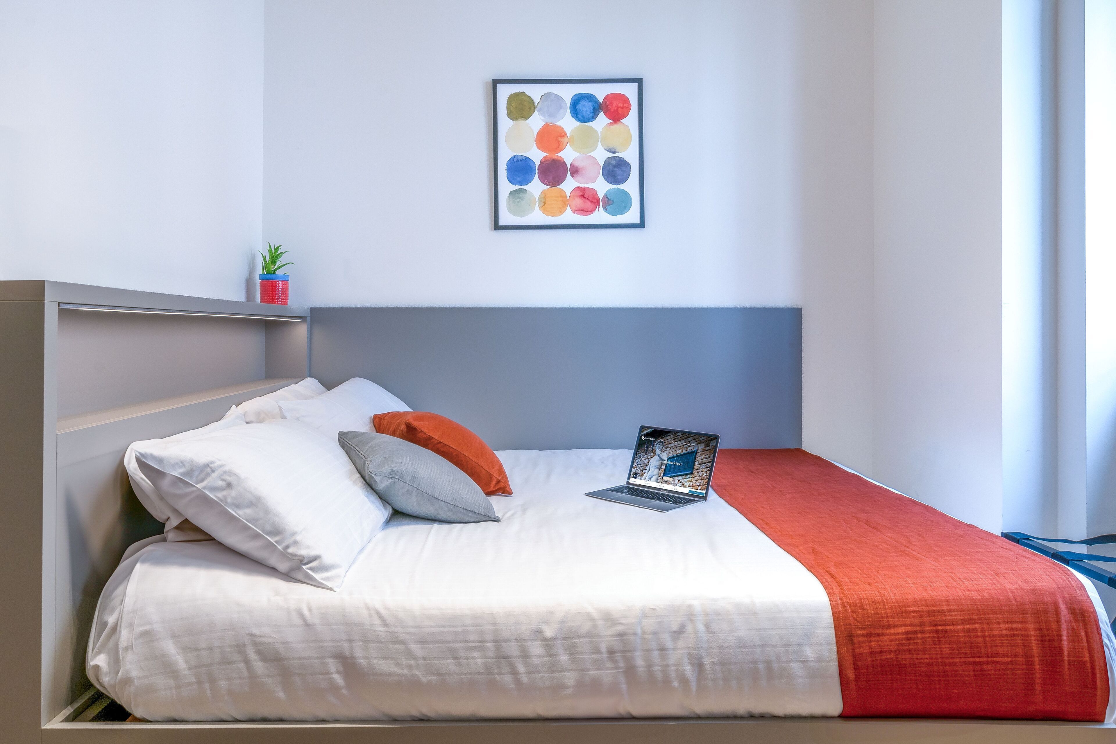 Un lit bien fait, avec une literie blanche et rouge, un ordinateur portable posé sur le lit et un tableau abstrait aux couleurs vives accroché au mur.