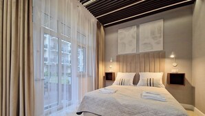 A modern bedroom with a large bed, two bedside tables, a headboard, and a view of the outside through a large window. - Baltic Home (Swinoujscie)