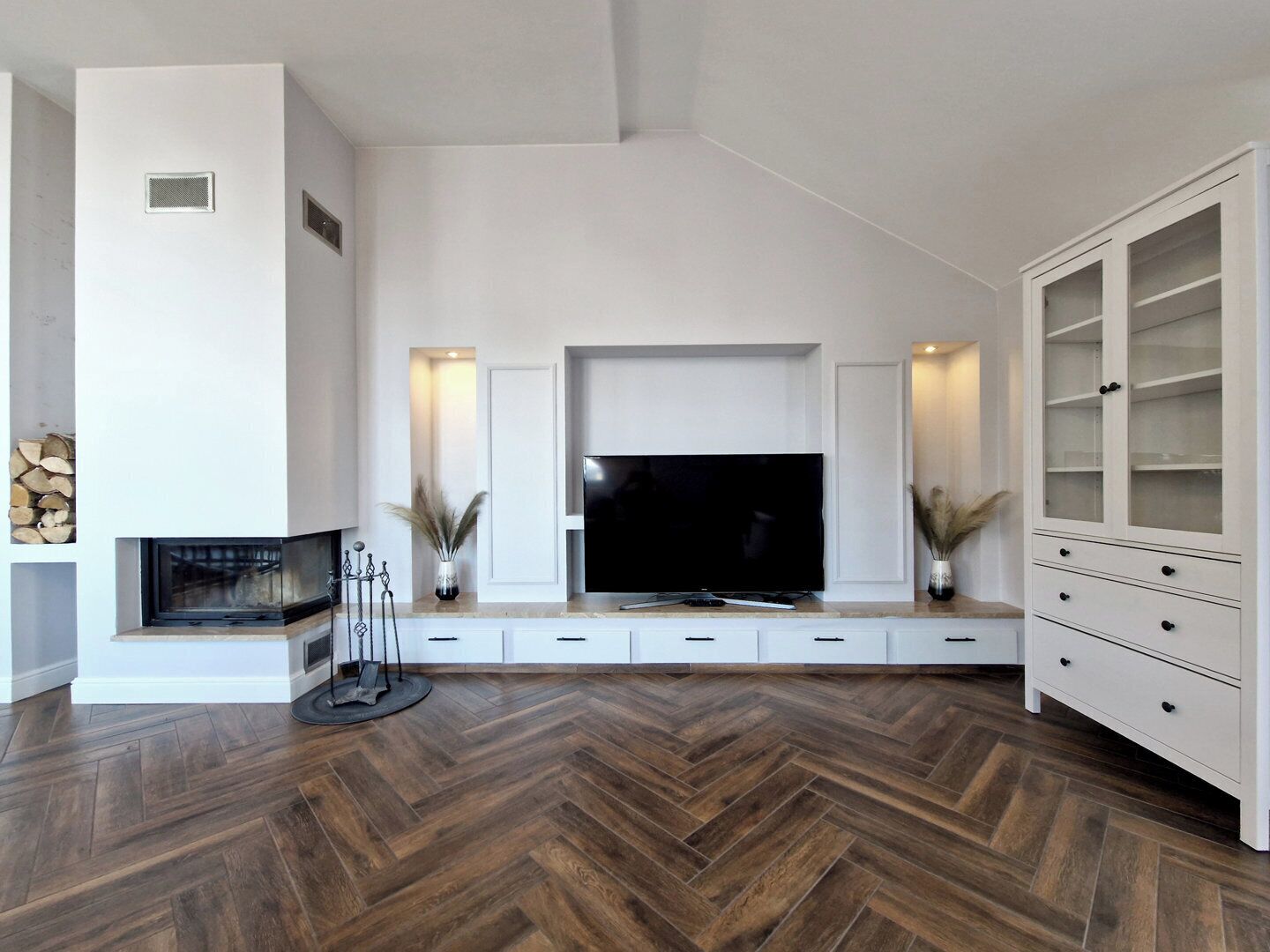 Uma sala de estar moderna com uma grande TV de tela plana, estantes embutidas e piso de madeira em padrão herringbone.
