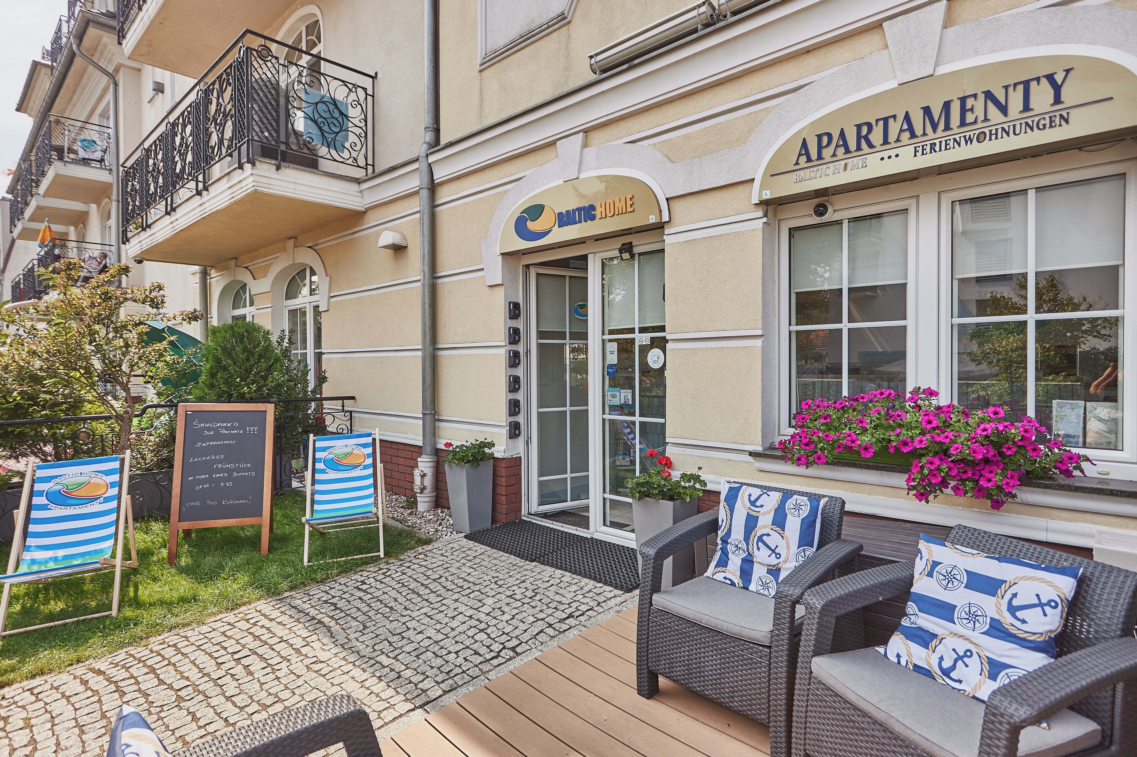 An exterior of a building with a sign reading "APARTMENTY" and "FELIENWOHNUNGEN". There are outdoor seating areas with wicker furniture and cushions, a small garden with flowers, and a paved entrance area.