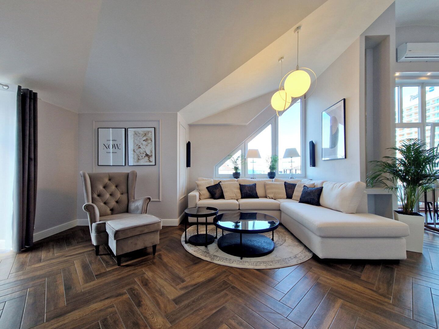 A modern living room with a herringbone wood floor, a tufted armchair, a white sectional sofa, and a round coffee table.
