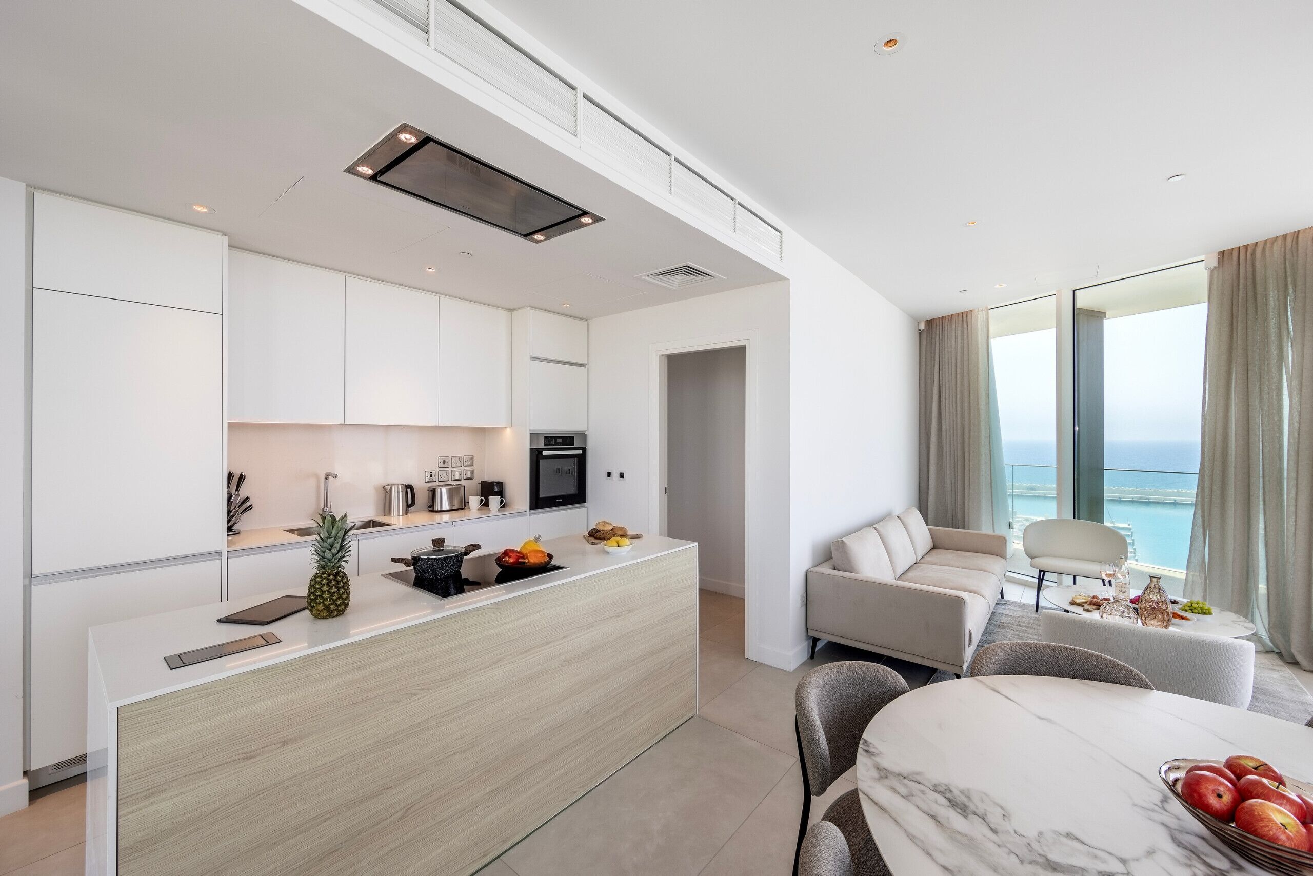 A modern kitchen with white cabinets, a central island with fruits, and a dining area with a marble table.