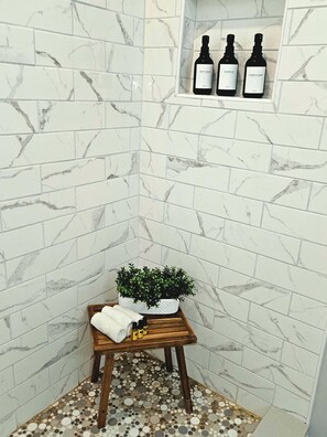 A bathroom with white marble tiles, a wooden stool with toiletries, and a shelf with three bottles.