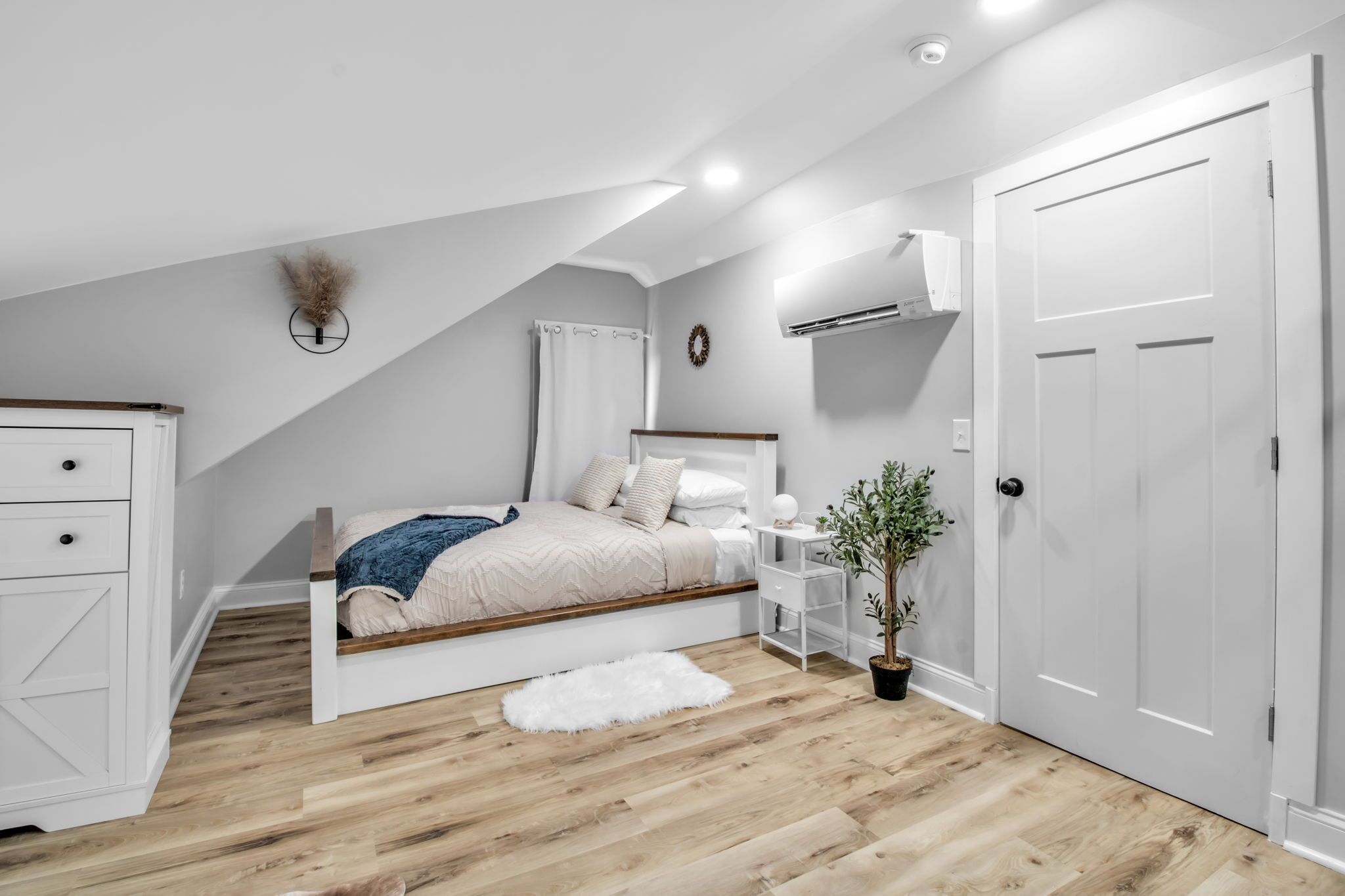 Une chambre avec un plafond en pente, un lit avec une tête de lit blanche, une table de chevet avec une lampe, une plante en pot et une porte blanche.