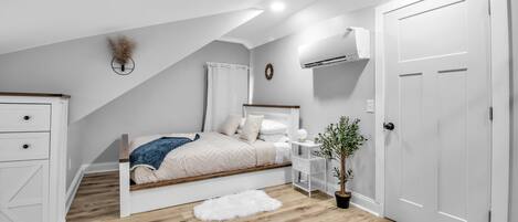 Une chambre avec un plafond en pente, un lit avec une tête de lit blanche, une table de chevet avec une lampe, une plante en pot et une porte blanche.