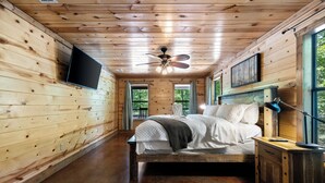 A wooden cabin bedroom with a bed, a ceiling fan, a mounted TV, and a window with curtains.