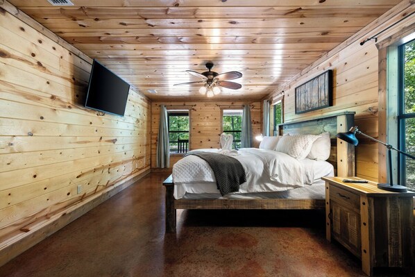 A wooden cabin bedroom with a bed, a ceiling fan, a mounted TV, and a window with curtains. - Dream Big is an entertainer's delight, offering a grand living space with 2,600 sq ft indoors and extensive covered wrap-around decks to put you on a front row seat for nature viewing. (Broken Bow)