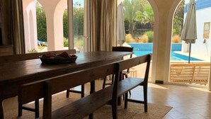 A dining area with a wooden table and benches, large windows with curtains, and a view of a swimming pool. - Charming Villa Son Parc (Son Parc)