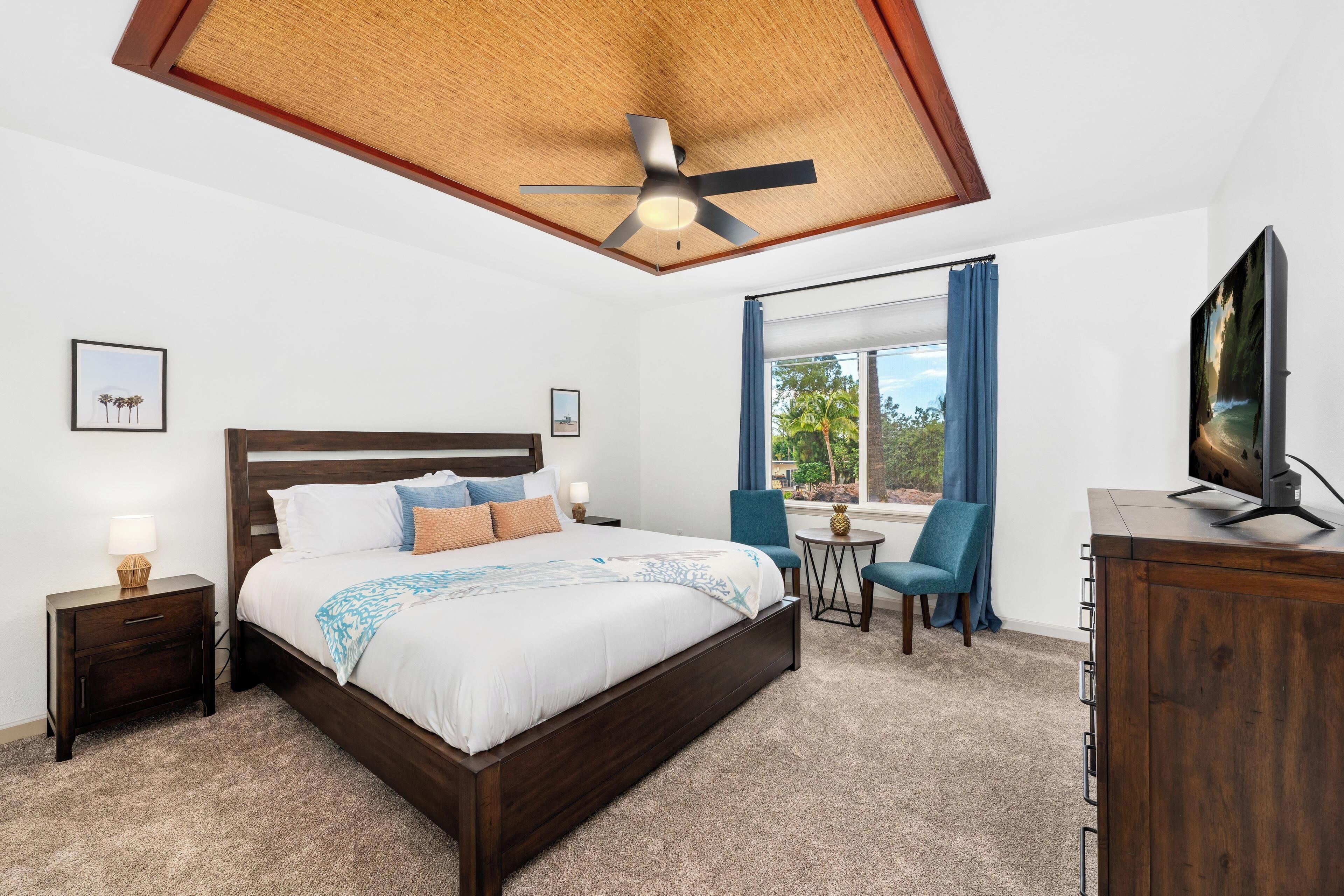Quarto de hotel com uma cama grande, um ventilador de teto, televisão, uma mesa pequena com cadeiras e uma janela com cortinas.