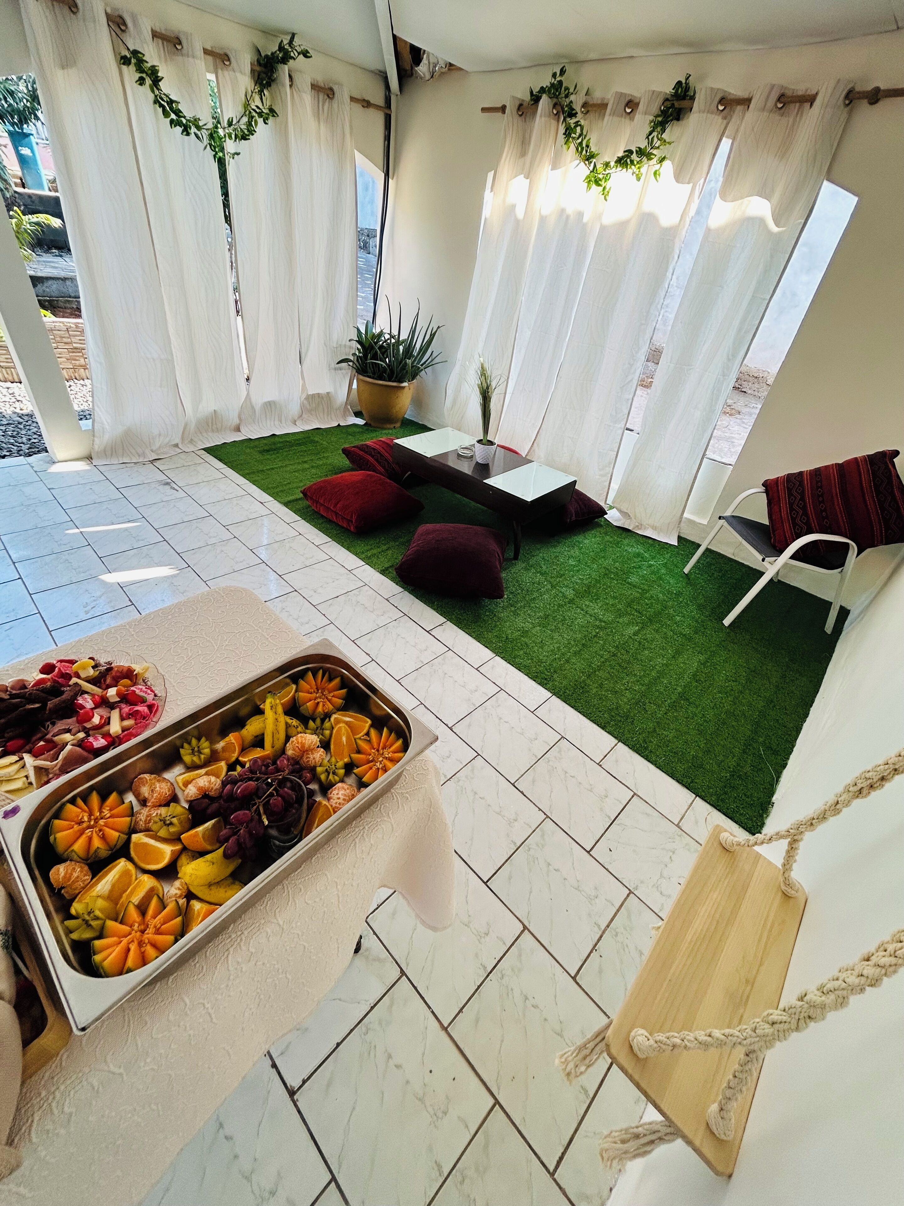 A room with a green artificial grass area, a table with a fruit platter, white curtains, and a hanging plant.