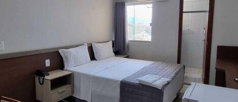 A hotel room with a bed, bedside table, chair, desk, and a window with curtains.