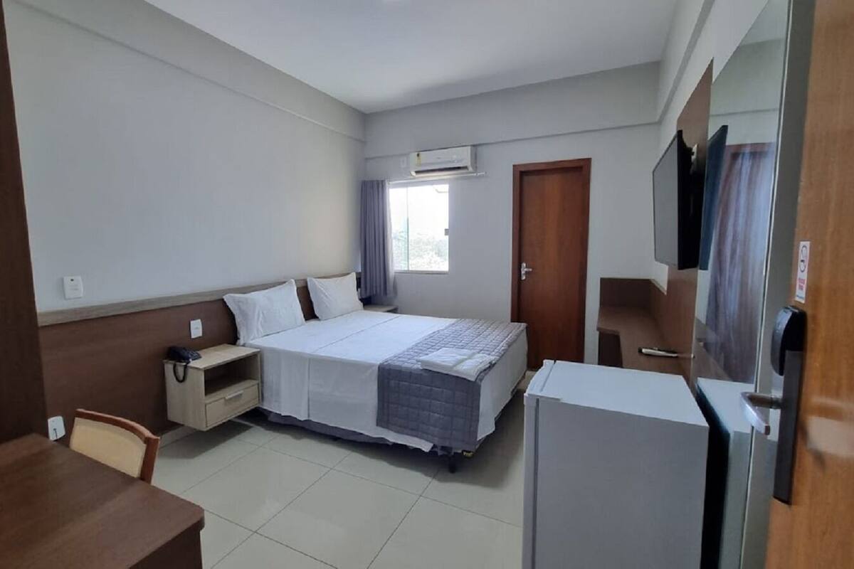 A hotel room with a bed, bedside table, chair, desk, and a refrigerator.