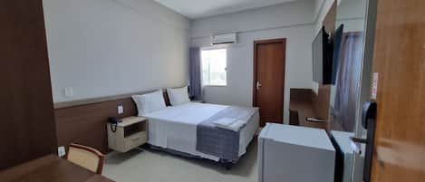 A hotel room with a bed, bedside table, chair, desk, and a refrigerator.