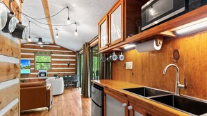Een houten interieur van een blokhut, met een kitchenette, een eethoek met tafel en stoelen, en uitzicht op groen buiten.