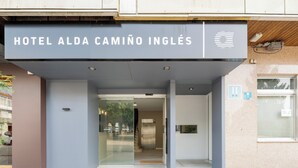 The entrance to Hotel Alda Camino Inglés with a clear glass door, a sign above the entrance, and a visible elevator. - Alda Camiño Inglés (Ordes)