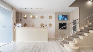 A modern reception area with a central desk, decorative wall art, a mounted television, and a staircase with a metal handrail. - Alda Camiño Inglés (Ordes)