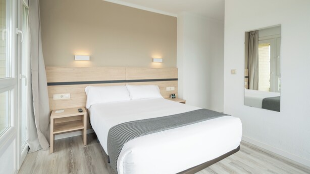 A hotel room with a large bed, bedside tables, a nightstand, a mirror, and a window with curtains. - Alda Camiño Inglés (Ordes)