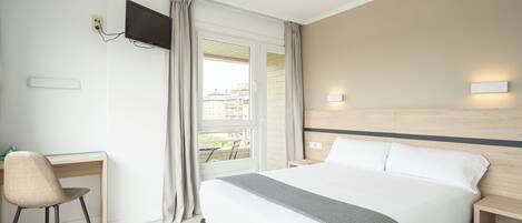 A hotel room with a large bed, a desk, a chair, a TV mounted on the wall, and a window with curtains.