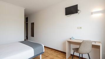 A hotel room with a bed, a desk, a chair, a TV mounted on the wall, and a light fixture above the desk.