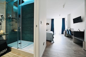 Una habitación de hotel moderna con cabina de ducha de vidrio, cama con cabecera gris, escritorio con silla y televisor de pantalla plana montado en la pared.