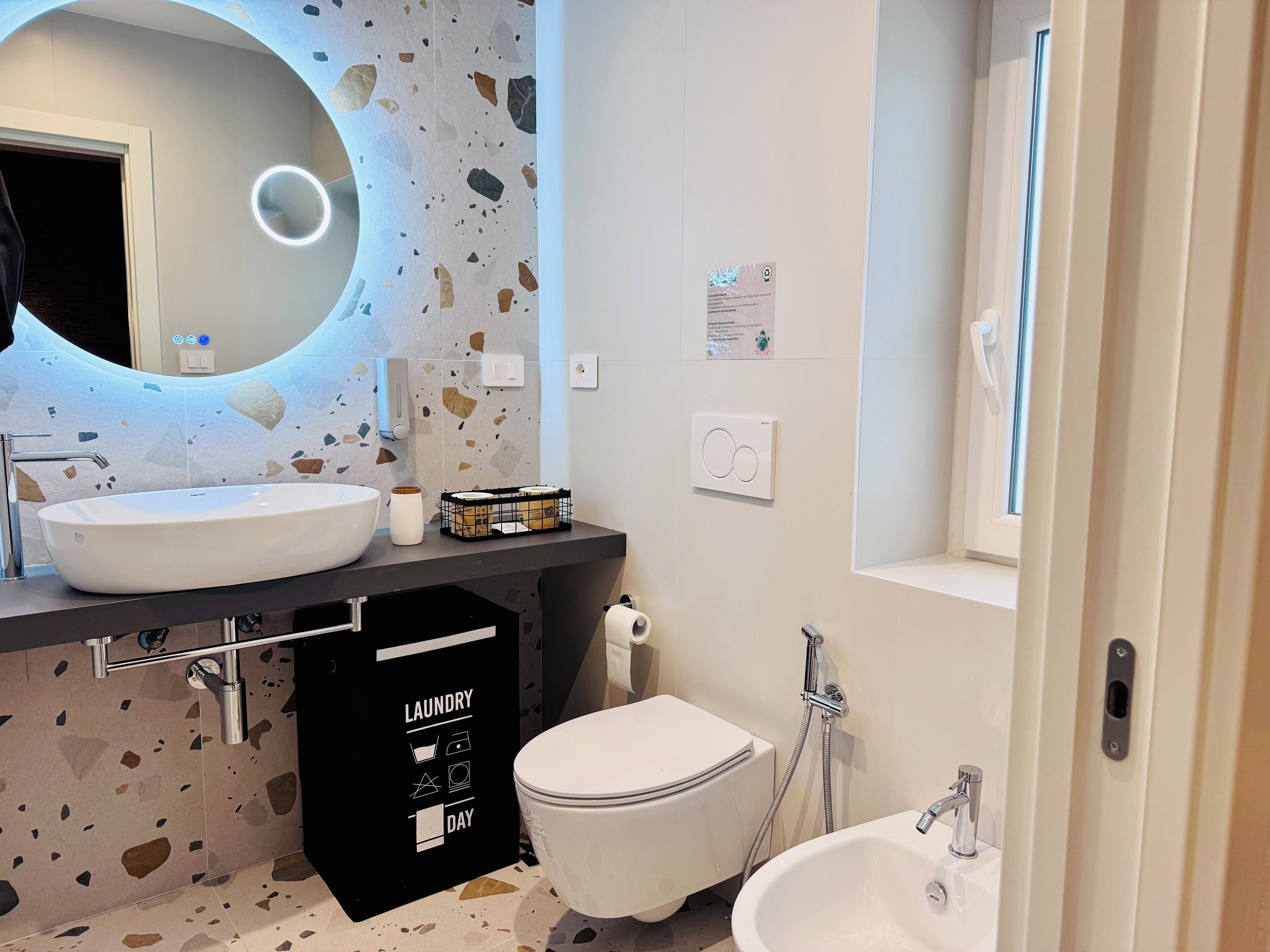 A modern bathroom with a large oval sink, a toilet, and a bidet. There is a black laundry basket with labels, a mirror with a circular light, and a window.