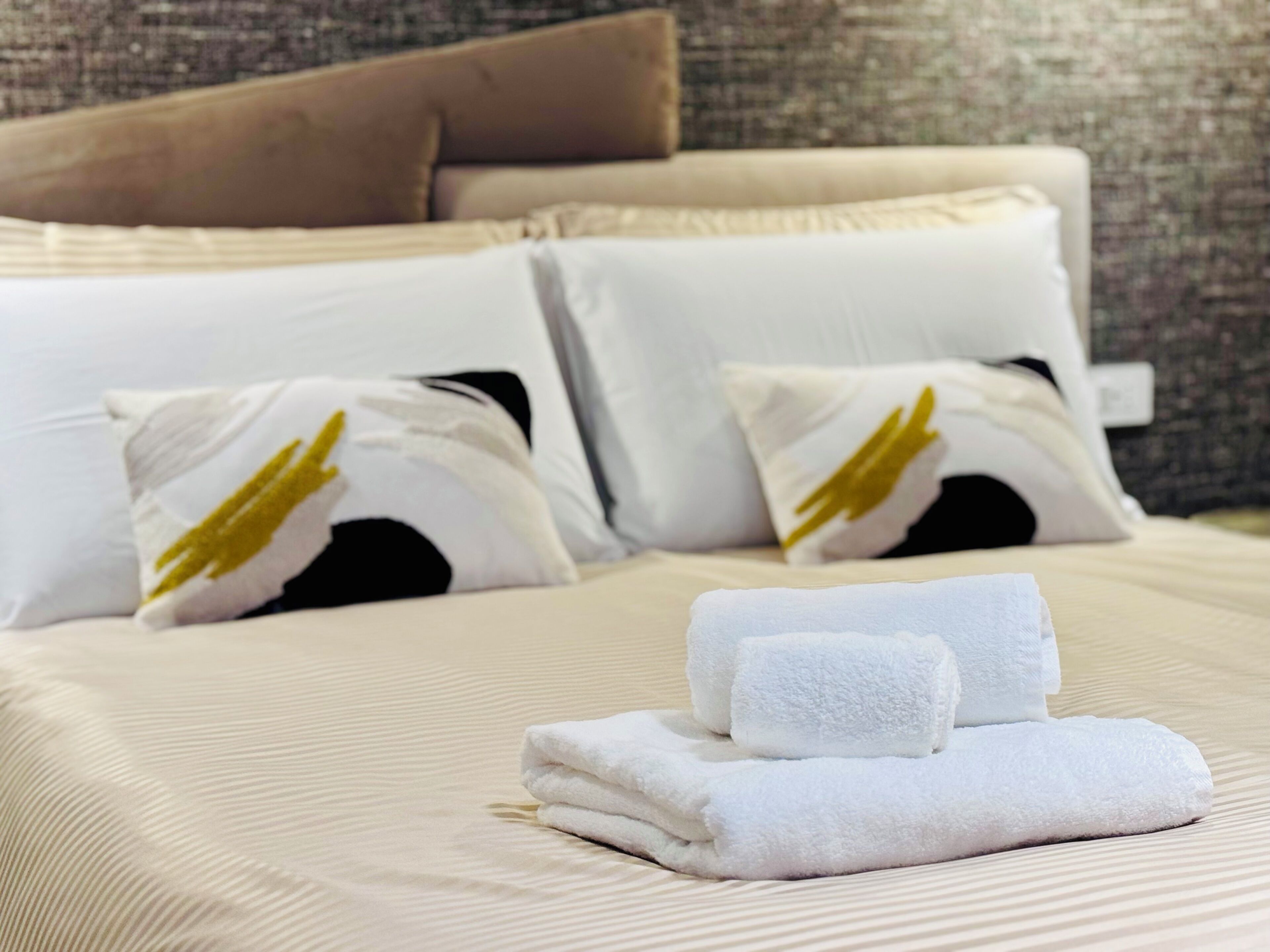 Una cama impecablemente hecha con sábanas blancas y dos almohadas con detalles dorados, una toalla blanca doblada y una pared texturizada al fondo.