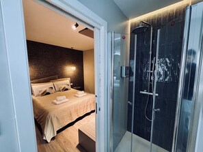 Quarto de hotel moderno com cama, chuveiro em cabine de vidro e parede texturizada.