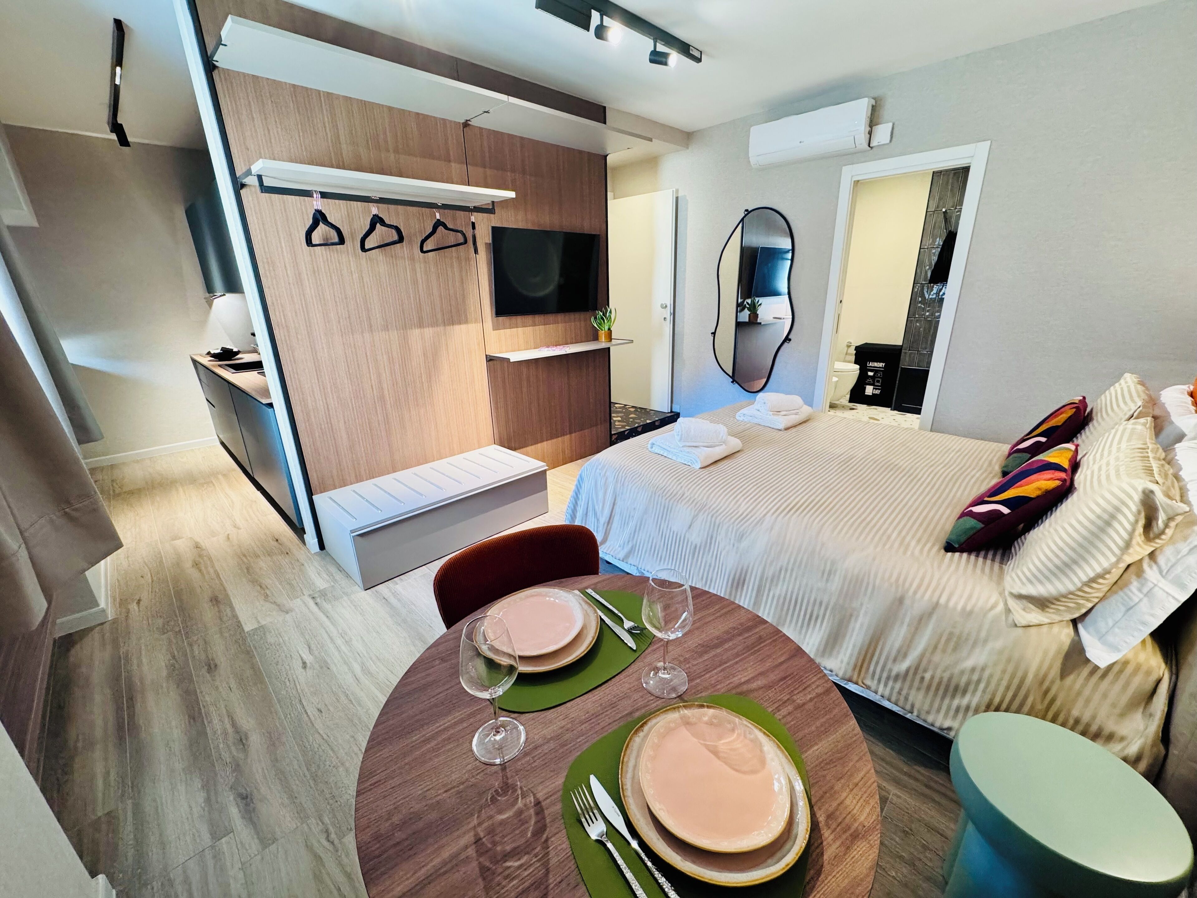 A modern hotel room with a bed, a dining area with a table set for two, a TV, and a mirror.
