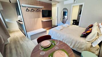 A modern hotel room with a bed, a dining area with a table set for two, a TV, and a mirror.