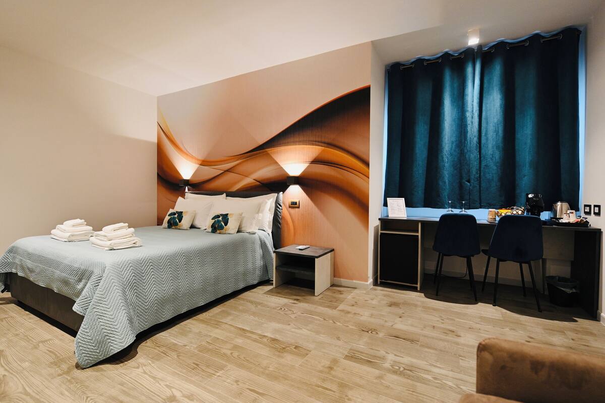 Habitación de hotel con cama, mesita de noche, escritorio, sillas y cortina.