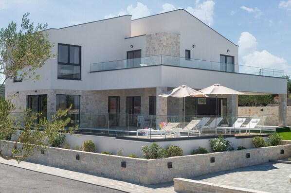 A modern two-story house with a balcony, stone facade, and outdoor seating area with umbrellas.