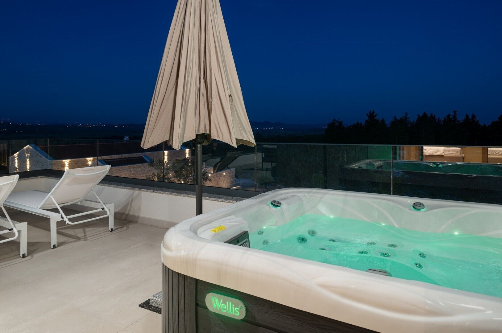 Un jacuzzi orné du logo « Wells », deux chaises longues blanches et un parasol sur une terrasse sur le toit, d’où l’on peut admirer la vue sur la ville au crépuscule.