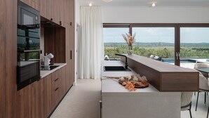A modern kitchen with a built-in oven, a large window, and a view of a pool and landscape. - Villa Terra & Aria Dalmatica pool sauna&jacuzzi (Sikovo)