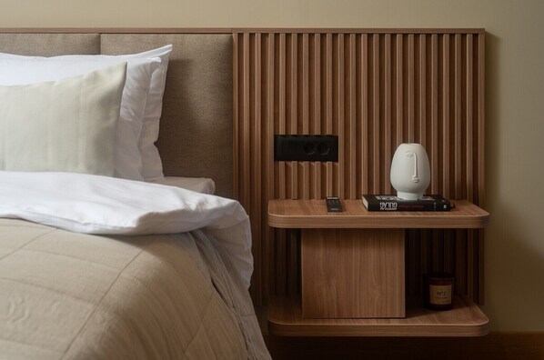 A neatly made bed with white linens, a wooden bedside table with a remote control, a book, and a candle.