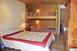 A wooden cabin room with bunk beds, a quilted bedspread, a nightstand with a lamp, and a wooden wardrobe.