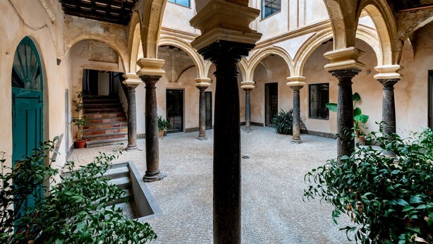 Un patio interior con arcos y columnas, una escalera y plantas en macetas.