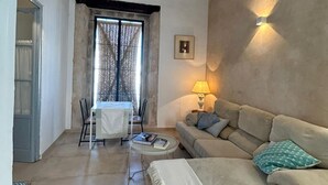 Una sala de estar con un sofá, una mesa y una lámpara. Hay una ventana con cortinas y una puerta que da a otra habitación.
