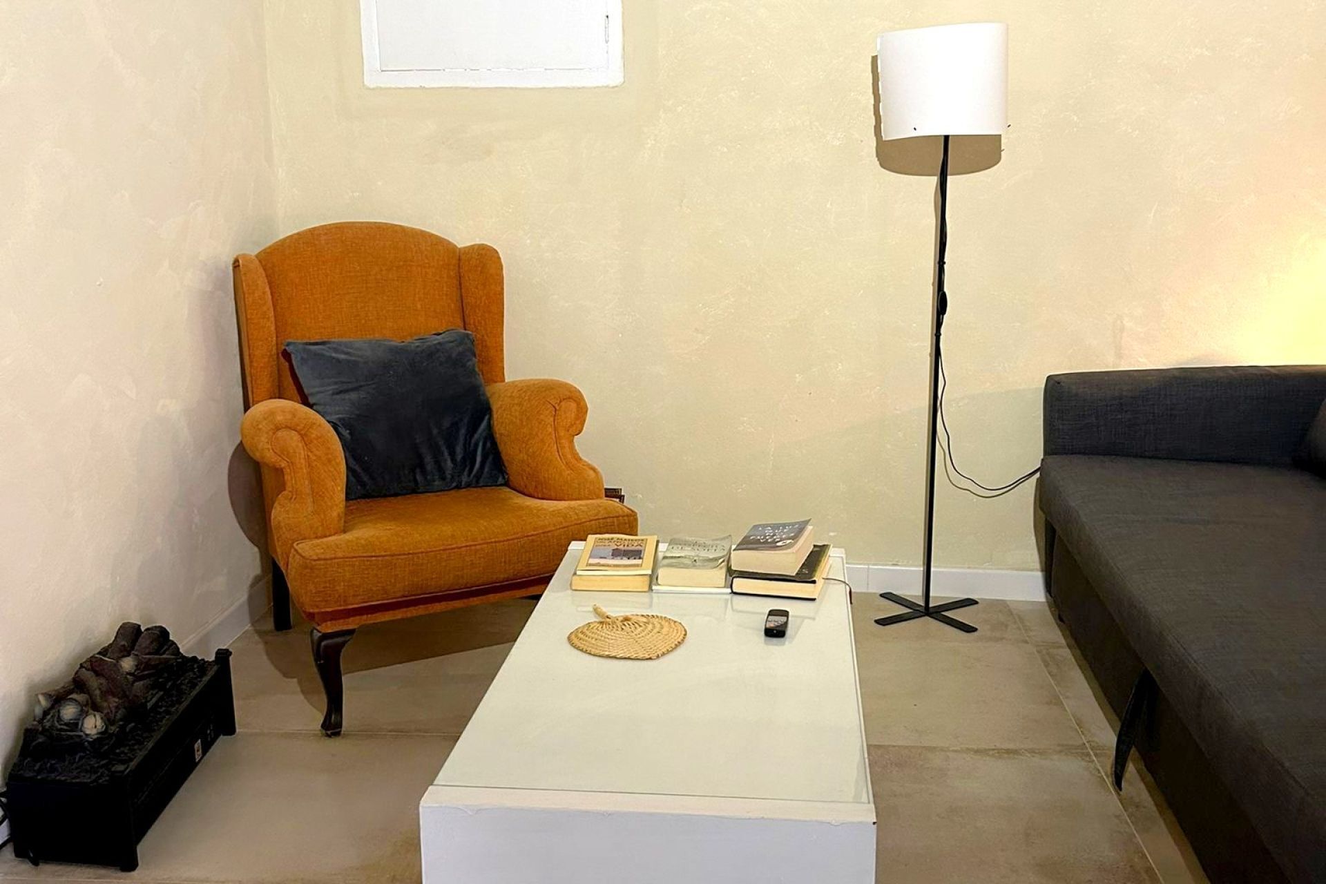 Uma sala de estar com uma mesa de centro branca, um sofá, uma poltrona laranja e um abajur.