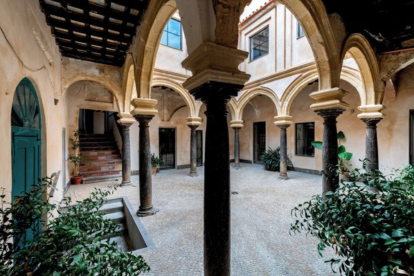 Stroll through the historic courtyard, surrounded by arches and columns, with a staircase leading to upper levels and lush greenery along the walkway.