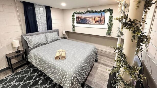 Un dormitorio moderno con una cama grande, una mesita de noche, un televisor montado en la pared y una planta decorativa.